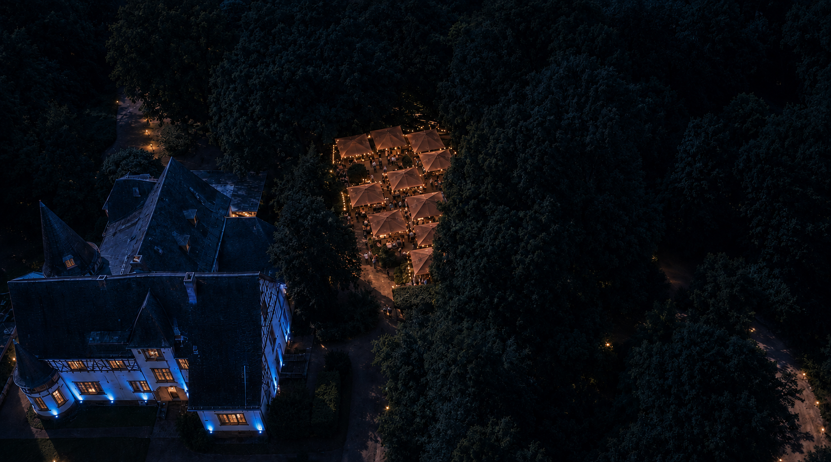 Abendliche Drohnenansicht des Oberwaldhauses mit beleuchteter Terrasse und Gästen unter großen Schirmen
